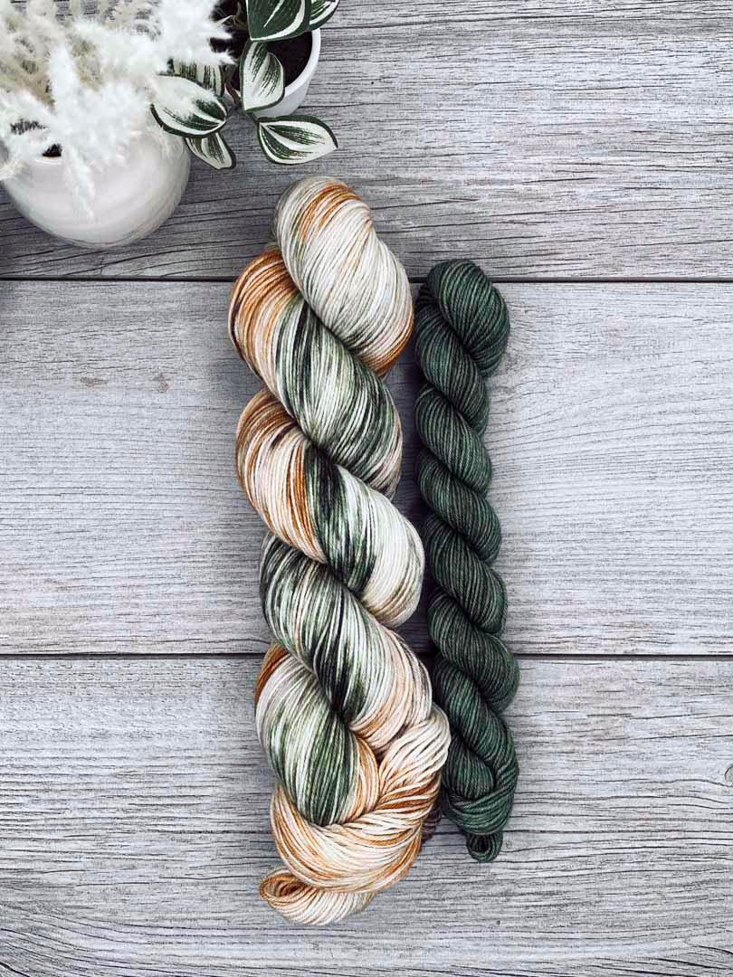 Three skeins of yarn in earthy tones on a wooden surface with a plant in the corner.