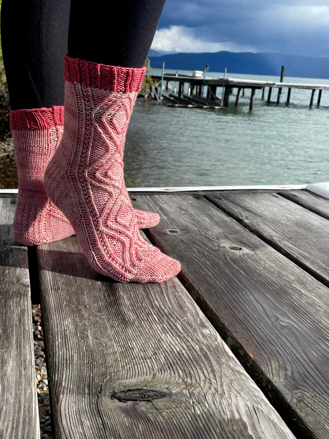 Pink knitted socks with red cuffs worn on a wooden dock by a body of water.