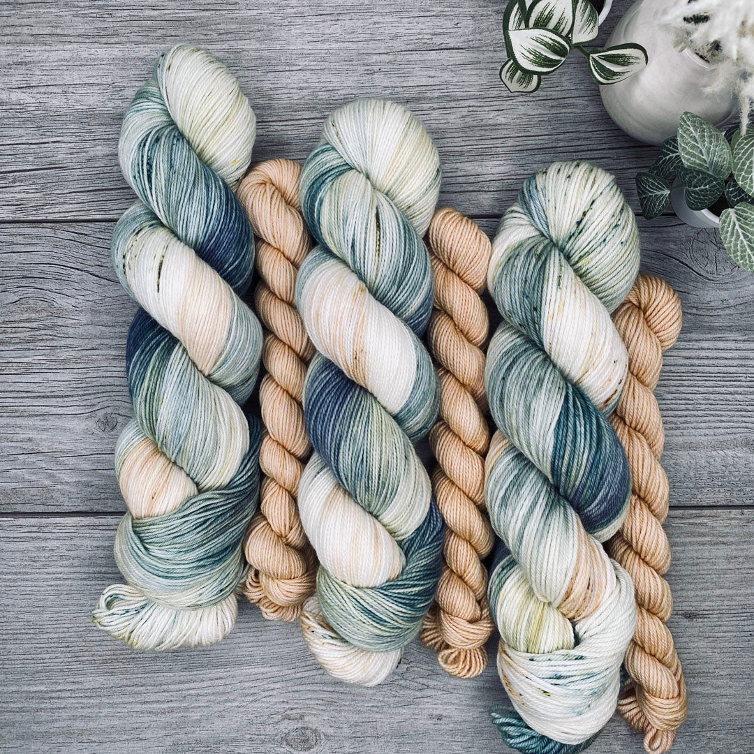 Three sock sets of variegated yarn laid flat on a gray wood surface, with the full skein in cream, teal, and blue-gray and the mini skein in pale butter yellow. Small potted plants appear in the top-right corner.