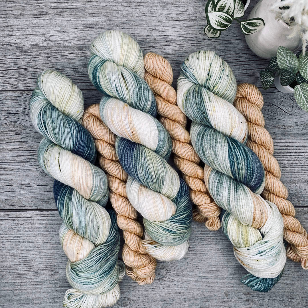 Three sock sets of variegated yarn laid flat on a gray wood surface, with the full skein in cream, teal, and blue-gray and the mini skein in pale butter yellow. Small potted plants appear in the top-right corner.
