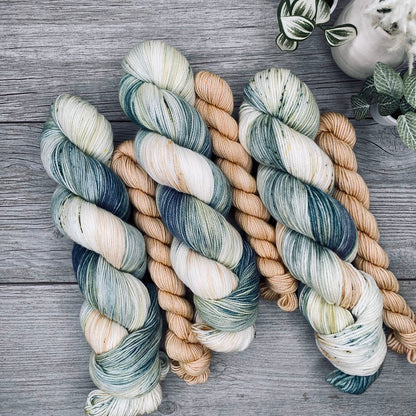 Three sock sets of variegated yarn laid flat on a gray wood surface, with the full skein in cream, teal, and blue-gray and the mini skein in pale butter yellow. Small potted plants appear in the top-right corner.