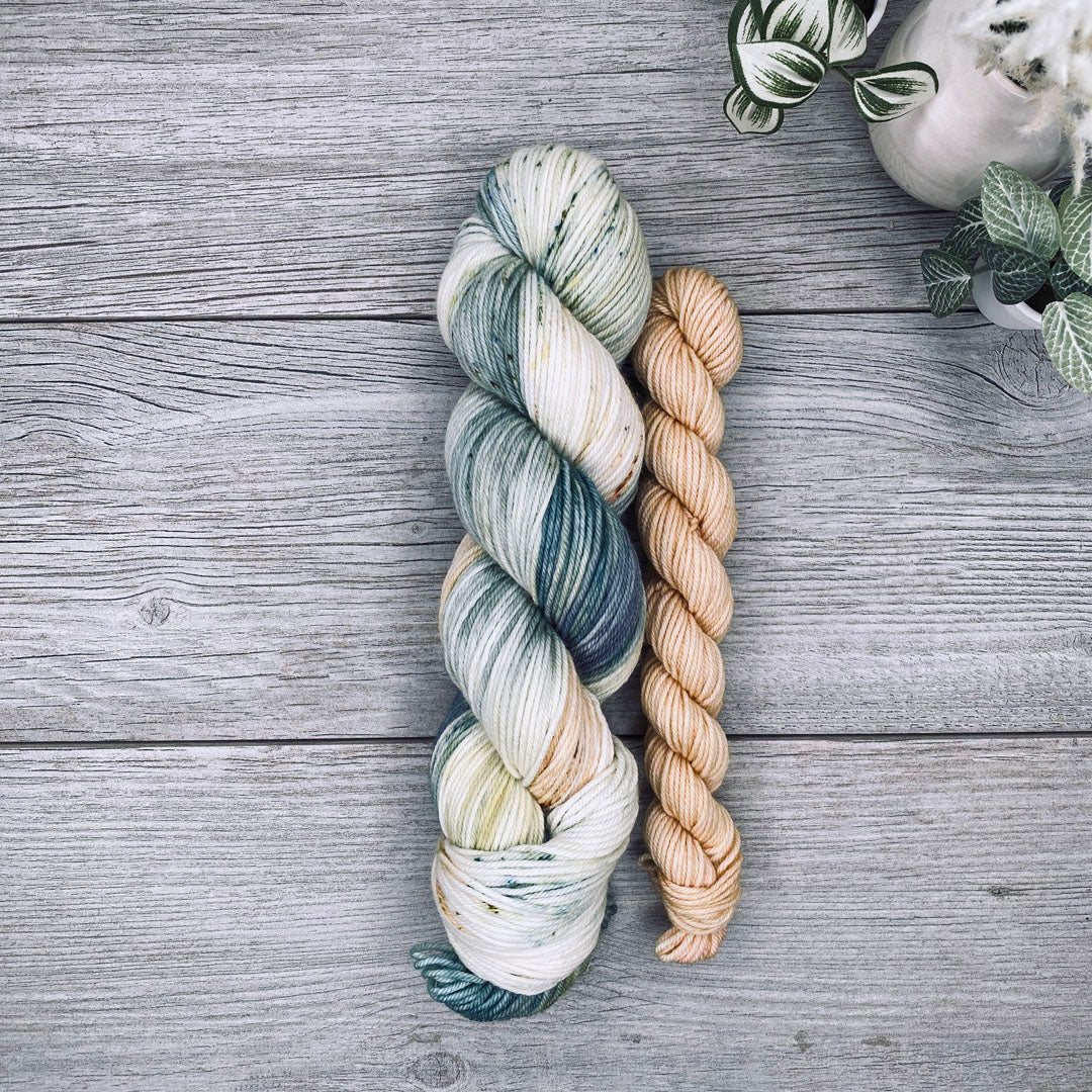 A sock set of variegated yarn laid flat on a gray wood surface, with the full skein in cream, teal, and blue-gray and the mini skein in pale butter yellow. Small potted plants appear in the top-right corner.