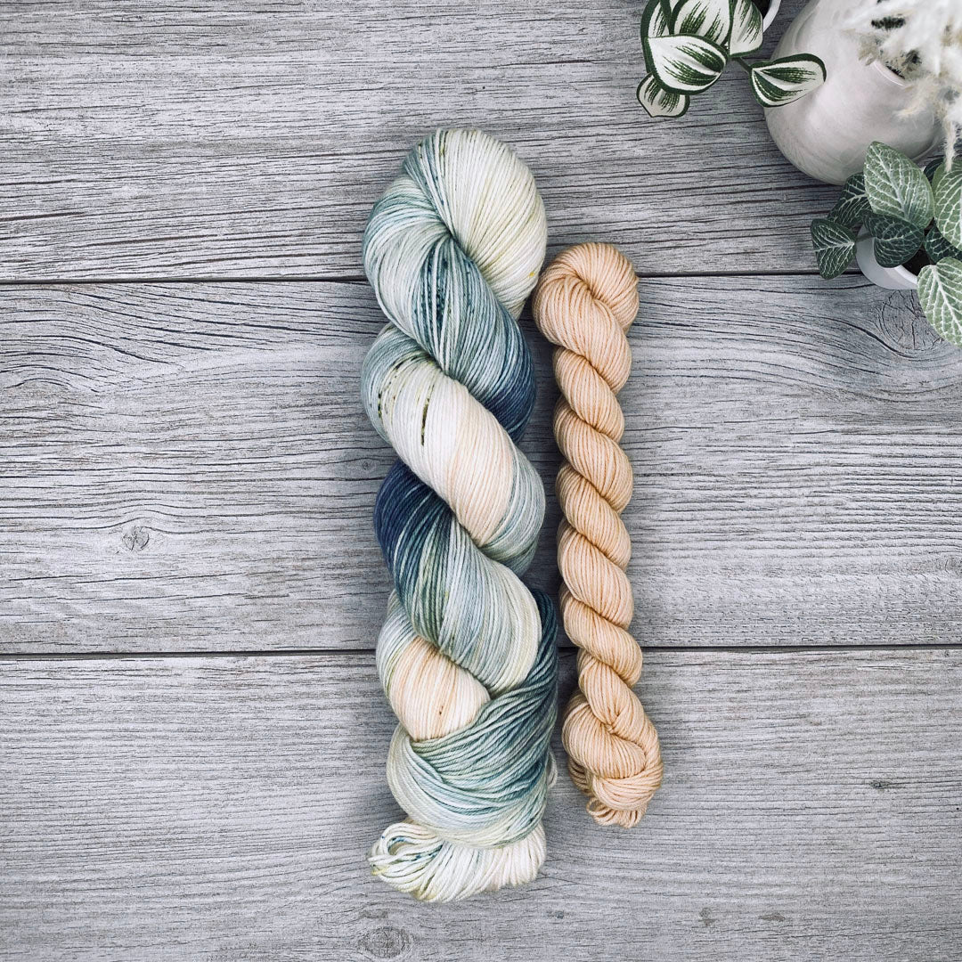 A sock set of variegated yarn laid flat on a gray wood surface, with the full skein in cream, teal, and blue-gray and the mini skein in pale butter yellow. Small potted plants appear in the top-right corner.