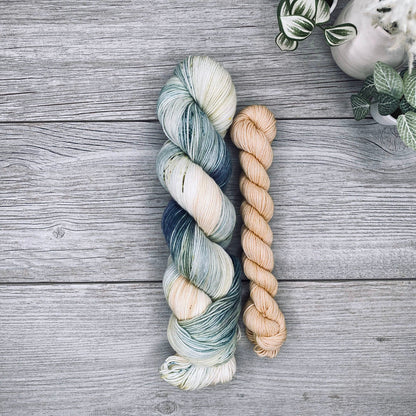 A sock set of variegated yarn laid flat on a gray wood surface, with the full skein in cream, teal, and blue-gray and the mini skein in pale butter yellow. Small potted plants appear in the top-right corner.