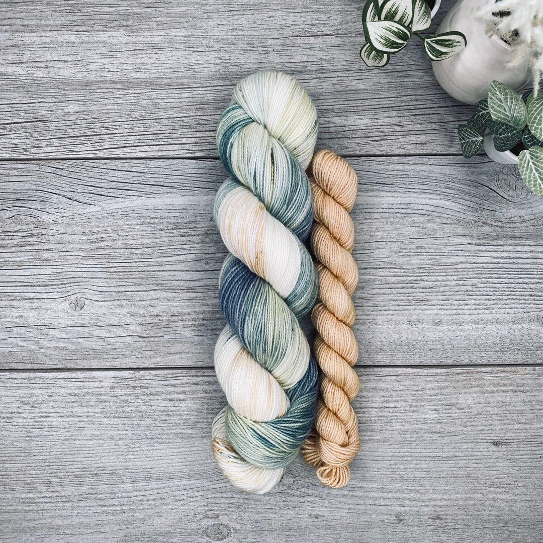 A sock set of variegated yarn laid flat on a gray wood surface, with the full skein in cream, teal, and blue-gray and the mini skein in pale butter yellow. Small potted plants appear in the top-right corner.