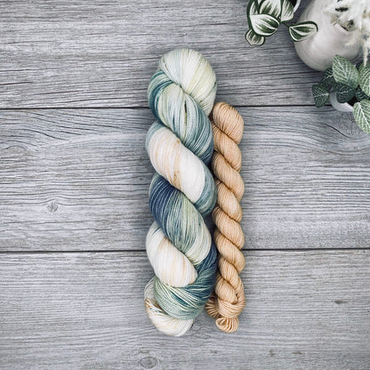 A sock set of variegated yarn laid flat on a gray wood surface, with the full skein in cream, teal, and blue-gray and the mini skein in pale butter yellow. Small potted plants appear in the top-right corner.